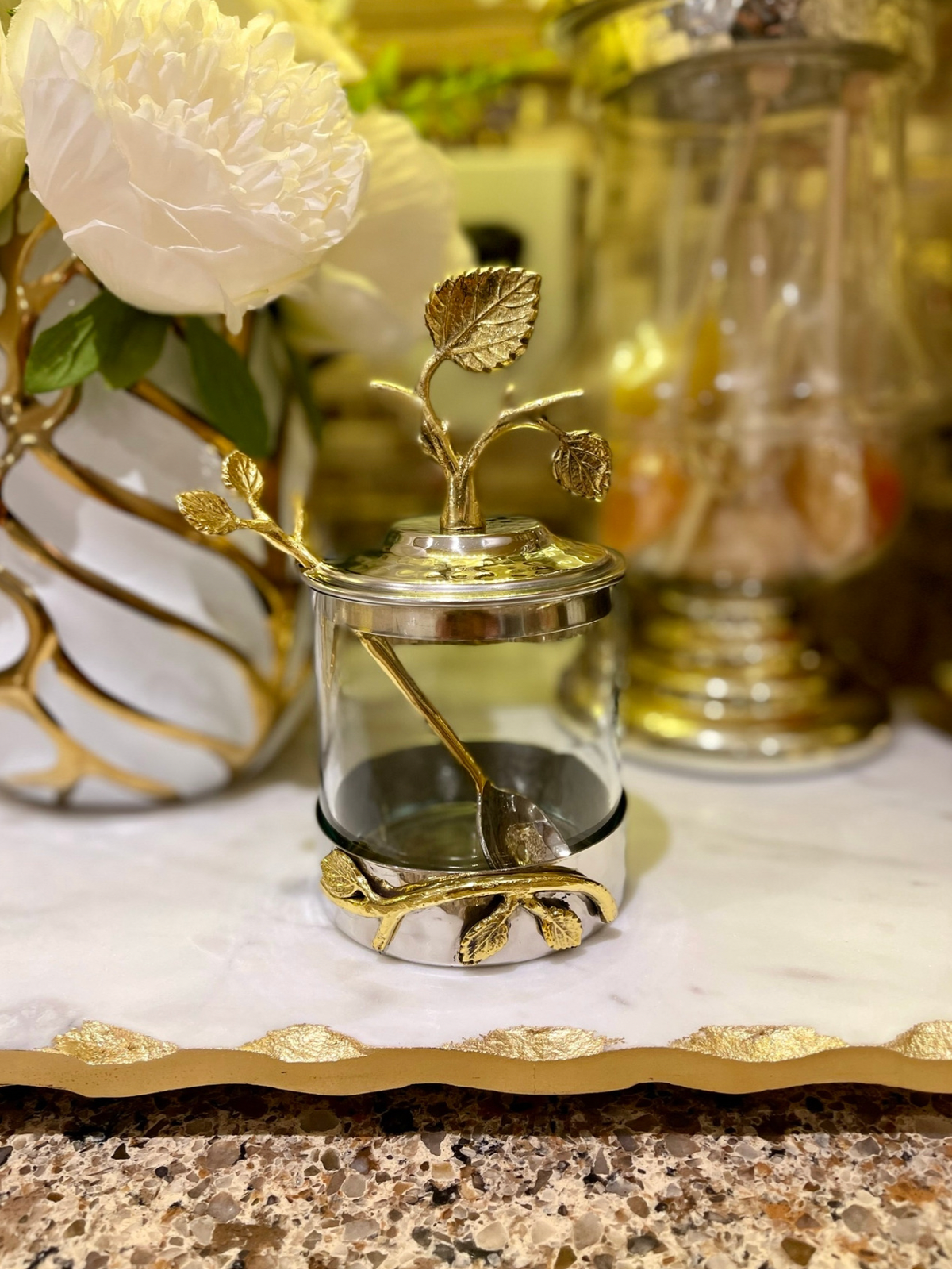 Bring vintage-inspired style to your kitchen or dining room table with this distinctive jar with spoon. It’s made of brass and features a silver-tone circular base and a metal lid with gold-tone raised stems, leaves, and flowers. 